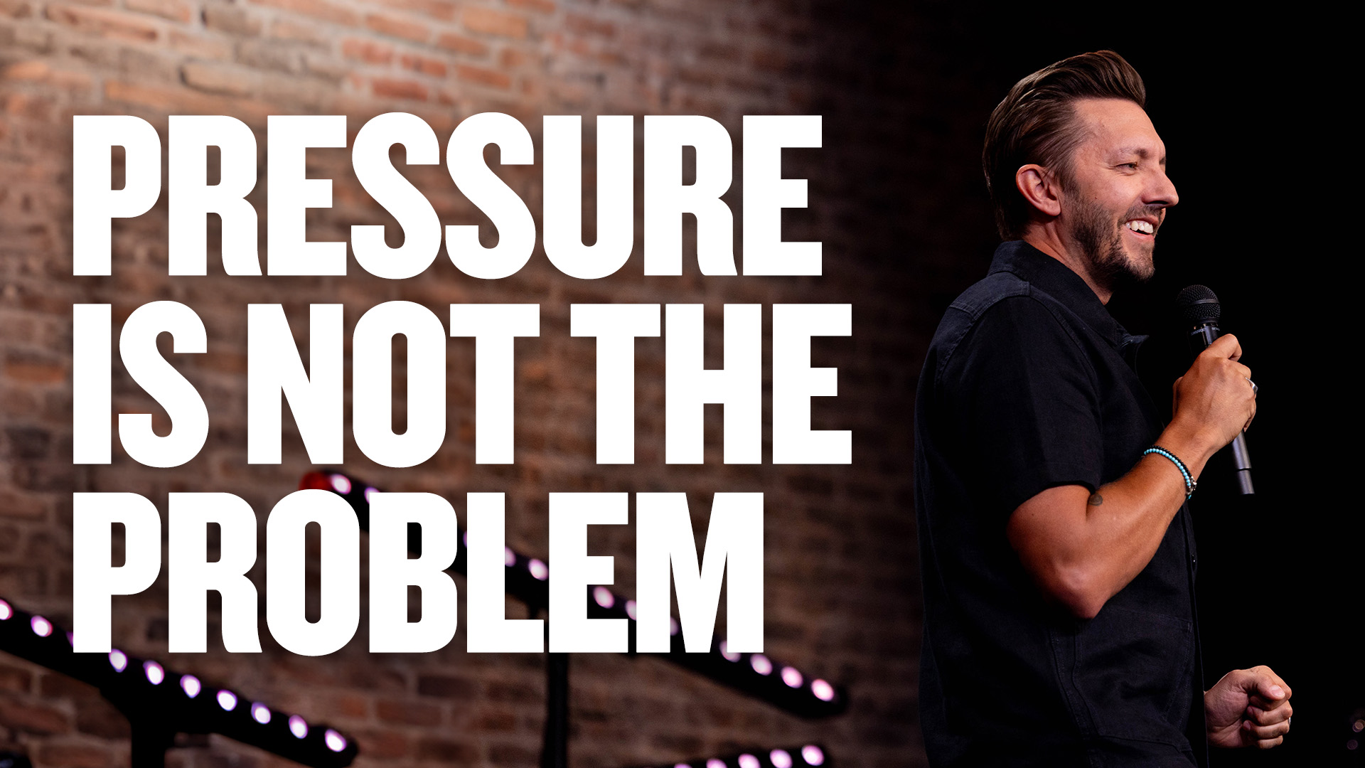 Pastor Levi Luskos stands on stage smiling holding a microphone up to his mouth with his right hand.